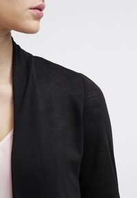 Schwarze Strickjacke mit weicher Textur, offener Vorderseite und langen Ärmeln. Kombiniert mit einem hellrosa Oberteil darunter.