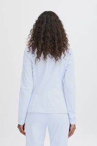 Žena s dugom kovrčavom kosom, u svijetloplavom odijelu, stoji okrenuta leđima, na jednostavnoj bijeloj pozadini.