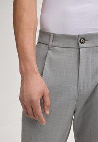 Graue maßgeschneiderte Hose aus strukturiertem Stoff, mit Falten und einem Knopfverschluss. Eine Hand befindet sich in der Nähe des Bundes.