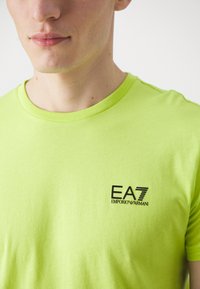 Neonově zelené bavlněné tričko s kulatým výstřihem a krátkým rukávem. Obsahuje černé logo EA7 Emporio Armani na levé části hrudi.