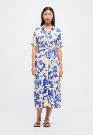 Vrouw draagt een beige midi-jurk met blauwe bloemenprint, korte mouwen, kraag, riem in de taille en zwarte sandalen met open teen, staand tegen een effen achtergrond.