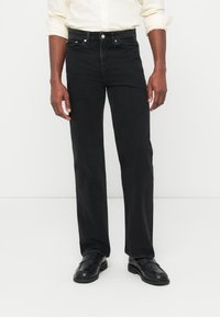 Hombre con vaqueros negros de pierna recta, mocasines de cuero negros y una camisa de manga larga color crema con las mangas remangadas, contra un fondo liso.