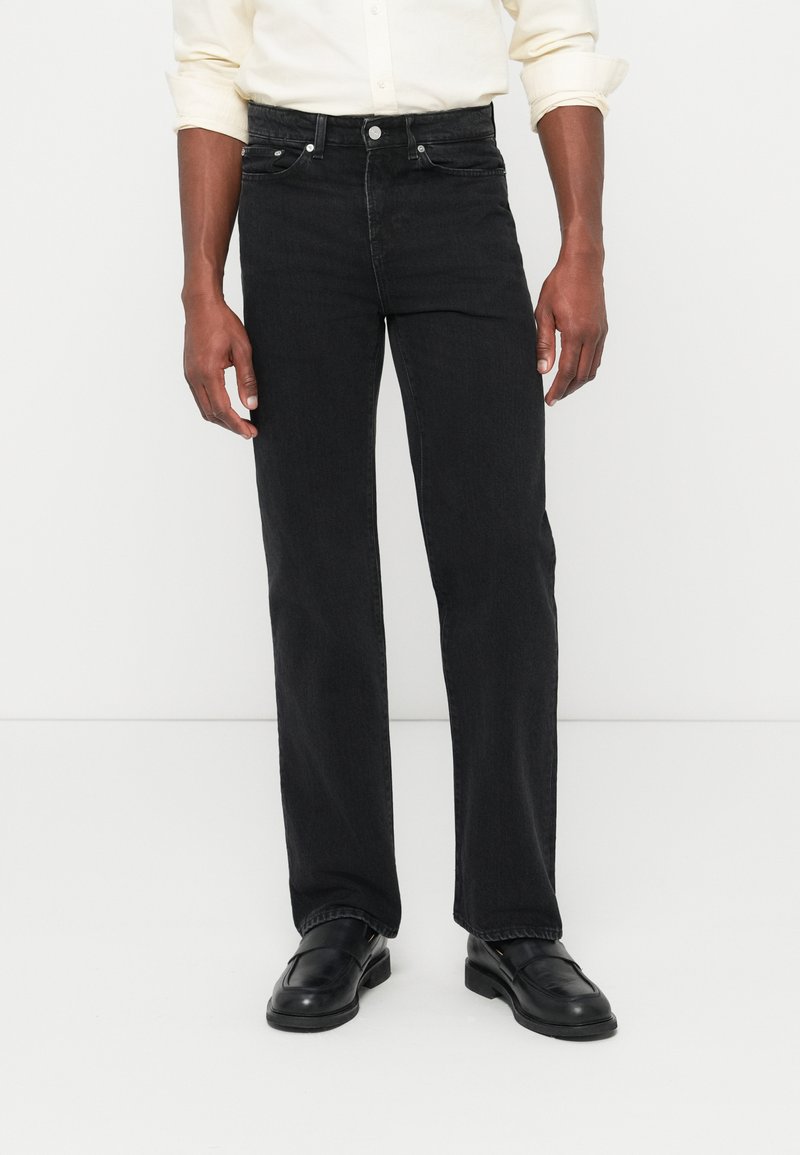 Hombre con vaqueros negros de pierna recta, mocasines de cuero negros y una camisa de manga larga color crema con las mangas remangadas, contra un fondo liso.