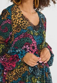 Femme portant une robe fleurie colorée avec bordure en dentelle et de grandes boucles d'oreilles en bois, mains reposant près de la taille.