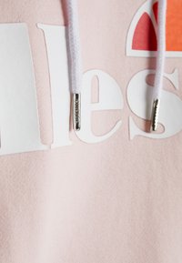 Sudadera de color rosa claro con texto blanco y extremos de cuerda plateados. Textura suave, diseño minimalista, que presenta un logo en colores vibrantes.