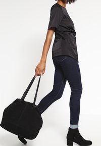 Černá taška přes rameno s měkkou sametovou texturou, dvojité černé úchyty a zaoblený tvar, nesena vedle tmavých džínů a kotníkových bot.