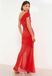 Czerwona wieczorowa suknia z dekoltem off-shoulder, marszczoną teksturą i przezroczystą dolną warstwą. Posiada dopasowaną sylwetkę i gładki materiał. Widoczne złote szpilki.