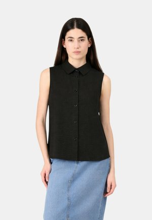 Donna con lunghi capelli scuri, che indossa una camicia nera senza maniche con bottoni e una gonna di jeans azzurro chiaro, in piedi contro uno sfondo semplice.