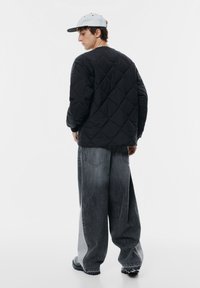 Sort gåsedunsjakke med diamantmønster, parret med brede mørkegrå jeans. Modellen bærer en lysegrå kasket. Minimalistisk design, ingen synlige logoer.