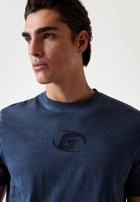 Mørkeblå t-shirt lavet af blødt stof, med et sort logo på brystet. Rund halsudskæring med korte ærmer og en let figursyet form.