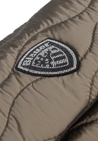 Nahaufnahme von gestepptem braunem Stoff mit einem schwarz-weißen dreieckigen Aufnäher, der kleine Perlendetails und stilisierten Text zeigt.