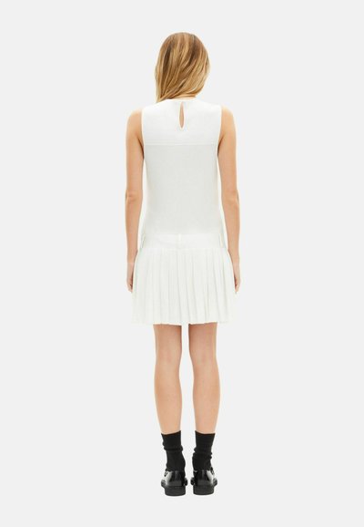 Vestito bianco senza maniche con gonna plissettata, dettaglio schiena a goccia e texture liscia. Indossato con calzini neri alla caviglia e scarpe.