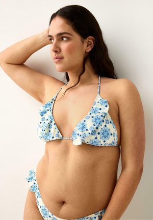 Kvinde med langt mørkt hår iført en hvid og blå bikini med blomsterprint og flæser, der ser til venstre med den ene arm hævet.