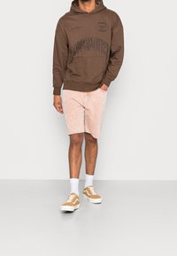 Felpa marrone con design grafico nero, abbinata a pantaloni corti di corduroy rosa chiaro e scarpe da ginnastica beige. Calzini bianchi visibili. Un outfit semplice e informale.
