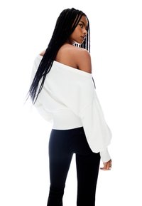 White oversized off-shoulder sweater with ribbed cuffs and hem, worn over black fitted pants, displayed on a model with long braided hair.