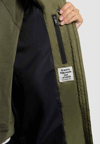 Interior de chaqueta verde oliva con forro negro; presenta un bolsillo con cremallera y una etiqueta de tela que dice "Un abrazo cálido hecho de tela."