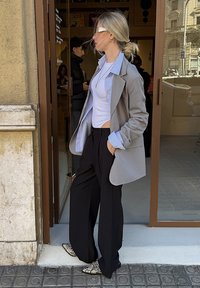Femme portant un blazer gris, un haut blanc, un pantalon noir et des chaussures en cuir de serpent, debout devant l'entrée d'un café en regardant sur le côté.