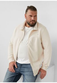 Homme avec une barbe et des cheveux attachés portant une veste zippée beige par-dessus un t-shirt blanc et un jean bleu, assis contre un fond blanc.