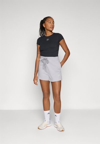Top nero a maniche corte con un piccolo logo, abbinato a pantaloni corti grigi chiari con vita regolabile e dettaglio verde del logo. Sneakers bianche e calze.