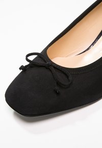 Ballerine plate en daim noir avec bout arrondi et petit noeud décoratif, dotée d'une doublure intérieure beige et d'un bord noir autour de l'ouverture.