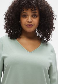 Νεαρή γυναίκα με σγουρά μαλλιά, που φοράει σκουλαρίκια με πέρλες και ανοιχτοπράσινο, υφασμάτινο τοπ με V-λαιμόκοψη, κοιτάζει μπροστά με ουδέτερη έκφραση.