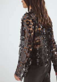 Musta paljettitakki, jossa on pyöreitä paljetteja, pitkät hihat ja avoin selkäosa. Kangas on sileä ja kiiltävä.