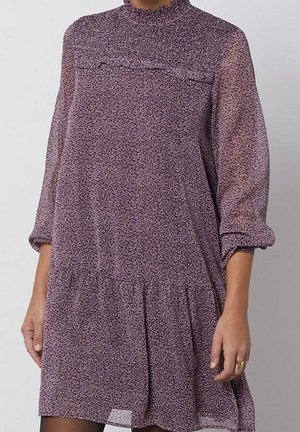 Kobieta ubrana w sukienkę z długim rękawem o fioletowym kwiatowym wzorze, z marszczonym, wysokim kołnierzem i talią.