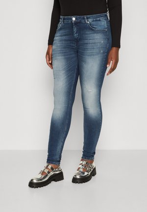 Osoba nosząca wyblakłe niebieskie obcisłe dżinsy i metaliczne srebrne buty na masywnej podeszwie z klamrami, stojąca na jednolitym tle.