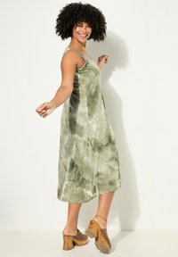 Grün gemustertes Tie-Dye-Kleid mit lockerer Passform, ärmellosem Design und knielanger Länge. Kombiniert mit Holzclogs mit goldenen Nieten.