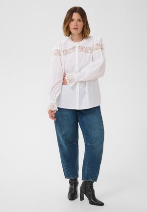 Femme portant un chemisier blanc à manches longues avec des détails en dentelle, un jean bleu et des bottines noires à talons hauts, debout devant un fond uni.