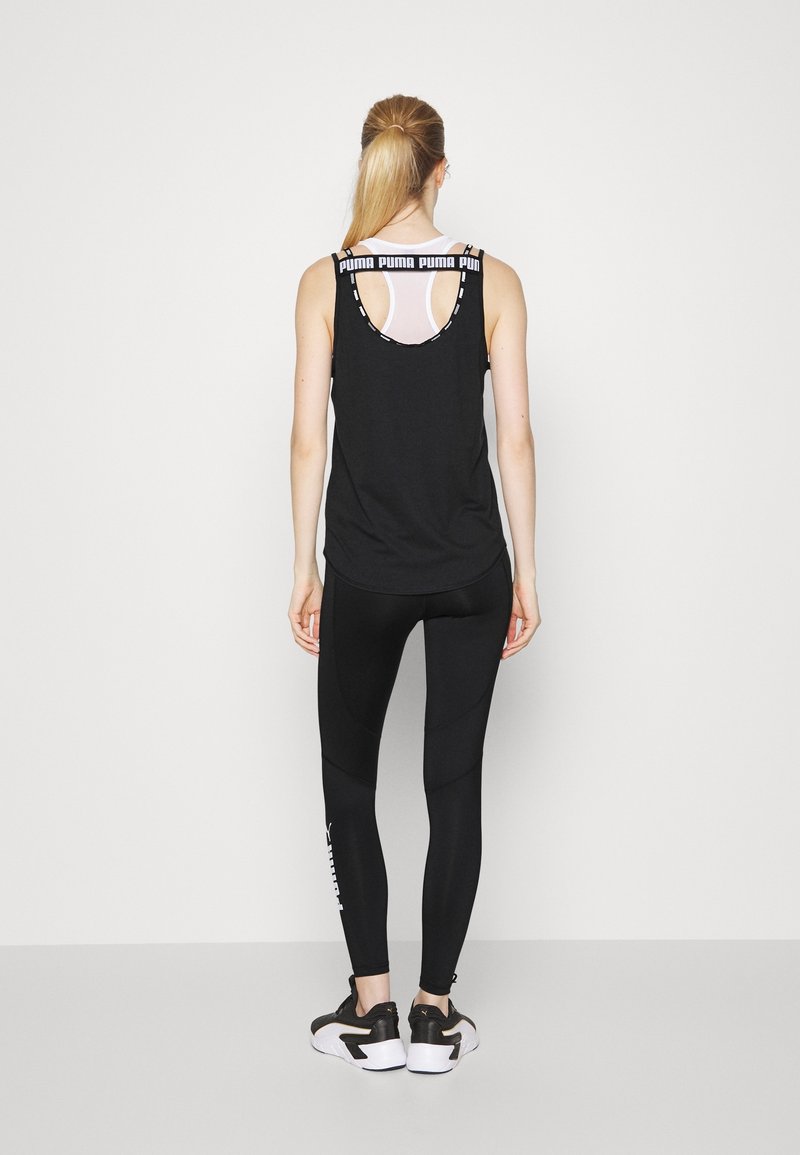 Femme avec une queue de cheval blonde portant un débardeur noir Puma, des leggings noirs avec un logo blanc, et des baskets noires et blanches, debout de dos.