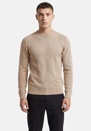 Mann trägt beige Strickpullover mit langen Ärmeln und schwarze Hose, steht vor einfachem hellem Hintergrund, schaut geradeaus mit neutralem Gesichtsausdruck.