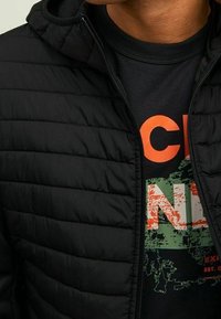 Chaqueta negra acolchada con textura acolchada y capucha, llevada sobre una camiseta negra con texto gráfico en colores naranja brillante y verde.