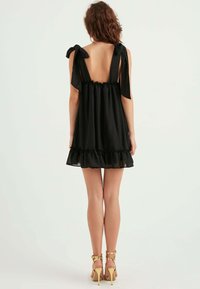 Schwarzes ärmelloses Kleid mit einem gerafften Saum, Rückenbindebefestigungen und transparenter Textur. Es hat ein offenes Rückendesign und goldene Riemchenabsätze.