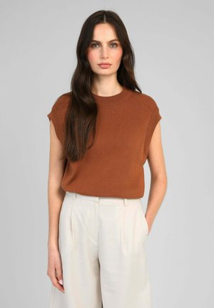 Femme aux longs cheveux foncés porte un pull sans manches couleur rouille et un pantalon blanc taille haute, debout devant un fond gris uni.