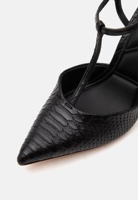 Chaussure femme noire à bout pointu avec motif texturé effet reptile et fine bride de cheville ornée d’un petit nœud en boucle.