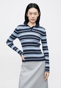 Πουλόβερ πόλο με ρίγες σε ναυτικό και ανοιχτό μπλε χρώμα, με ριμπ υφή και γιακά. Φορεμένο με γκρι φούστα, διαθέτει μακριά μανίκια και εφαρμοστή γραμμή.