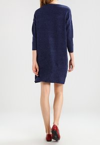 Navyblå stickad klänning med trekvartsärmar, lös passform och rundad halsringning, bar med röda skor som visar ben under knäet.