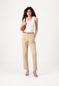 Calças beiges de cintura alta, corte ajustado, combinadas com uma blusa sem mangas branca com acabamento em renda. Cinto castanho e bolso, sandálias com tiras.