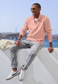 Light pink zip-up sweatshirt, gray jeans, and white sneakers with gray accents. Sunglasses reflect surrounding light; casual outdoor setting.