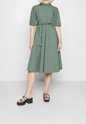 Kvinde iført en grøn kjole i mellemlang længde med puffede ærmer, et bundet taljebælte og sorte hæle med tynde stropper.