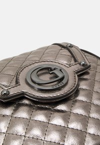 Syrad quiltad metallic grå handväska med en cirkulär svart logotyp, silverfärgade detaljer och en slät, strukturerad yta.