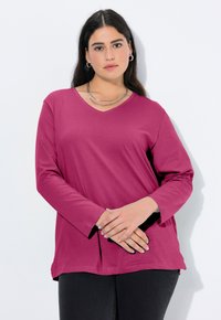 Bluzka z długim rękawem i dekoltem w serek w głębokim odcieniu magenty o luźnym kroju i bocznych rozcięciach; w zestawieniu z ciemnymi jeansami i warstwowymi srebrnymi naszyjnikami.
