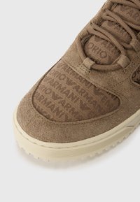 Zapatilla de ante marrón con parte superior texturizada que presenta patrones de logotipos impresos, punta redonda y suela de goma beige con diseño estriado.