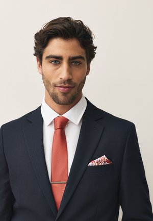 Man with dark hair and beard wearing navy suit, white shirt, orange textured tie, and patterned pocket square against a plain background.