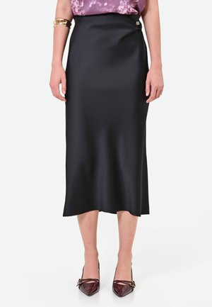 Femme portant une jupe midi noire, un chemisier violet à motifs, un bracelet doré et des talons pointus rouge foncé, debout sur fond blanc.