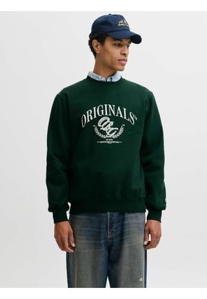Homme portant un sweat-shirt vert foncé avec le texte "ORIGINALS" et une casquette de baseball bleu marine, debout devant un fond clair uni.