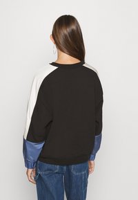 Sudadera negra con mangas de satén en contraste en crema y azul; cuello redondo, puños acanalados y ajuste relajado. Pantalones vaqueros visibles en la cintura.