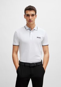Camisa polo blanca con cuello gris y ribete negro, que presenta un logo de "BOSS" en el pecho. Se lleva con pantalones y cinturón negros.