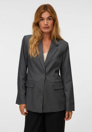 Femme aux longs cheveux blonds portant un blazer gris foncé à un seul bouton et un pantalon noir, debout devant un fond uni clair.